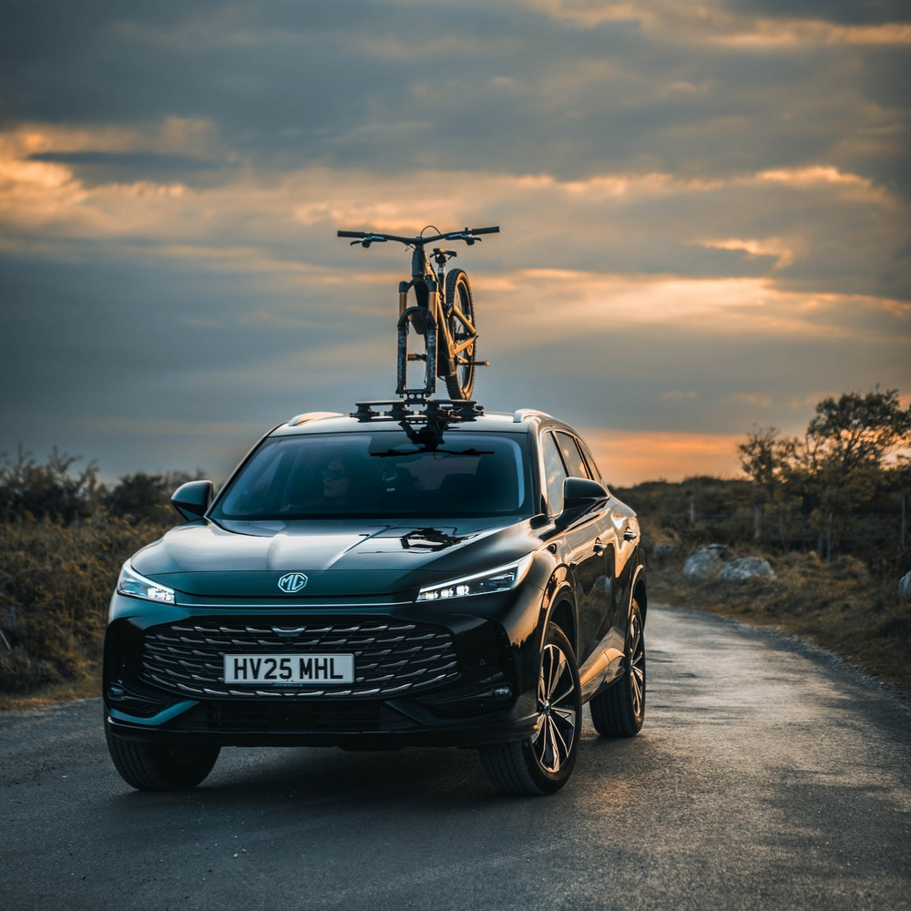 A dark metallic SUV parked on a road at sunset with a bicycle mounted on its roof rack. The vehicle has a large front grille, sleek headlights, and alloy wheels, with a license plate reading HV25 MHL. — view 9